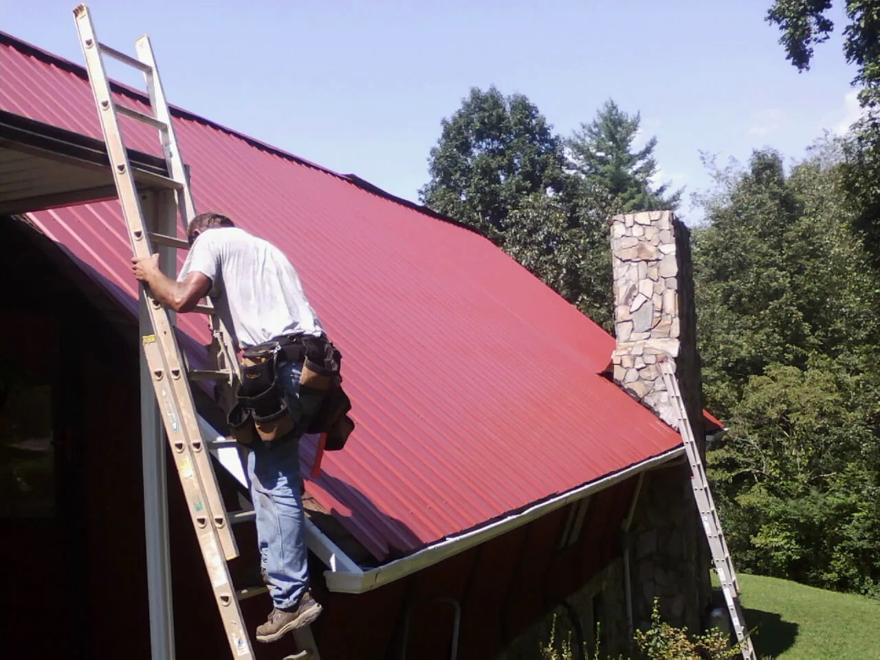Roofer on a ladder beside a red metal roof and stone chimney — Soffit & Fascia Repair by Roofing Brothers in Golden